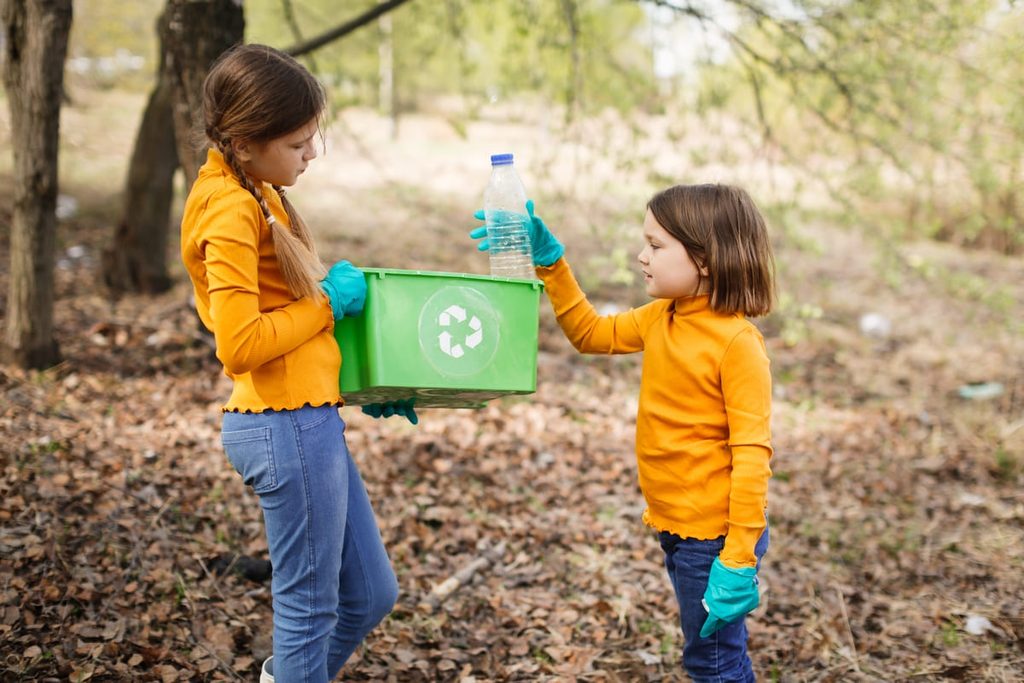 Dos niñas reciclando una botella de plástico en un contenedor verde durante una actividad de educación ambiental al aire libre.